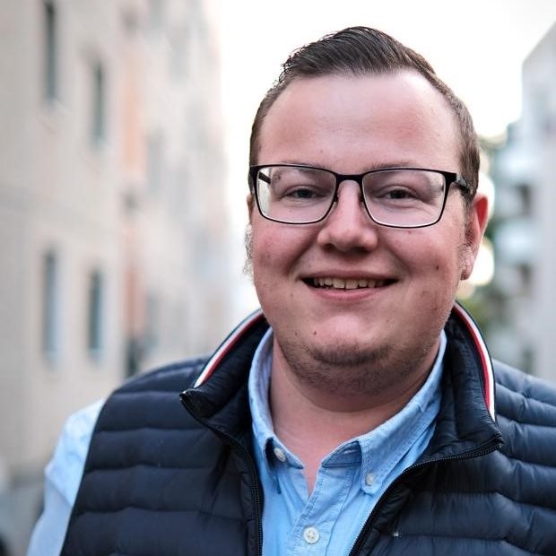 Lächelnder Mann mit Brille, dunkler Weste und hellblauem Hemd vor einem urbanen Hintergrund.